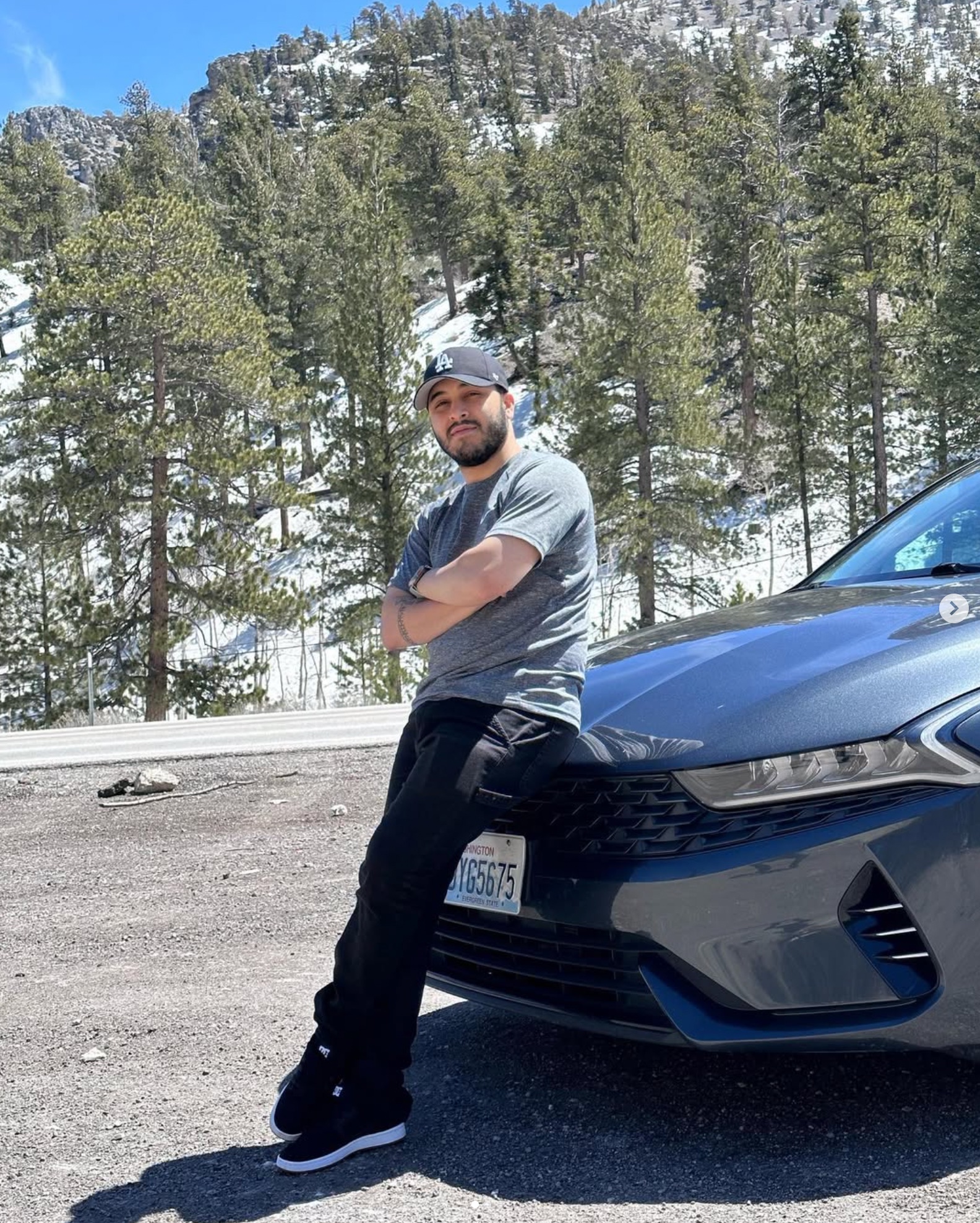 Eddryni Winddston de boné e braços cruzados, ao lado de um carro em uma paisagem de montanhas com neve.